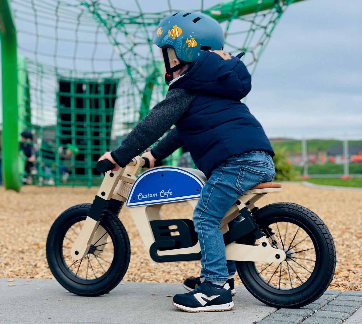 A child wearing a blue helmet and navy puffer jacket rides a wooden balance bike with a blue and white 'Custom Cafe' frame on a paved playground path, with a large green climbing net in the background.