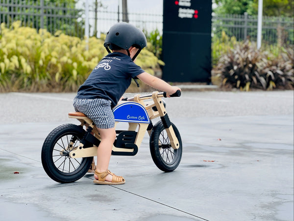 A child wearing a black helmet and dark t-shirt rides a wooden balance bike with a blue fuel-can style tank on a paved outdoor area.